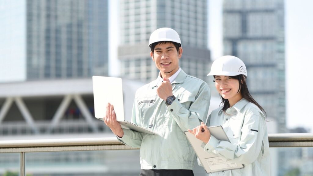Two Asian construction engineers, a male and a female, smiling confidently with a modern cityscape in the background. The man is holding a laptop, and the woman is taking notes, both wearing white safety helmets and work attire, representing professional urban development and teamwork in the construction industry.